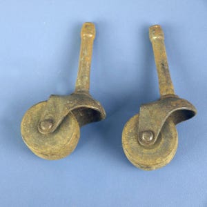 Może przedstawiać: Dwa antyczne metalowe kółka zardzewiałe. Każde kółko ma małe koło, zakrzywiony wspornik i długi trzpień. Koła są okrągłe i wydają się być wykonane z twardego materiału. Kółka są prawdopodobnie do mebli.