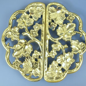 May include: A gold-colored, openwork metal decorative piece, possibly a trivet or serving accessory. The design features a symmetrical arrangement of stylized leaves and flowers, with a central vertical split. The piece has a rounded, quatrefoil shape.