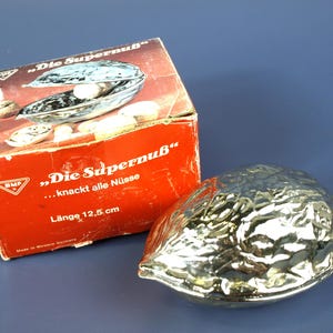 以下が含まれることがあります： クルミの形をした銀色の金属製くるみ割り器と、オリジナルの赤い段ボール箱。箱には「Die Supernuß」および「knackt alle Nüsse」という文字と、12.5cmという印字された長さが記載されています。くるみ割り器の表面は質感のある仕上がりです。