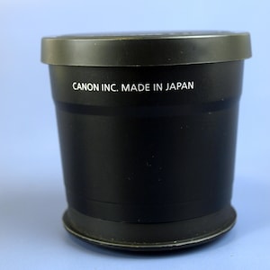 May include: A black Canon lens cap with the text "CANON INC. MADE IN JAPAN" in white. The cap is cylindrical with a flat top and a slightly wider base. The background is a light blue.