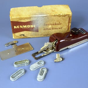 May include: A vintage Kenmore buttonhole attachment set. The set includes a maroon and silver buttonhole machine, a metal plate, and several gray plastic buttonhole templates. The original box is included, with the Kenmore logo and "buttonhole attachment" printed on it.