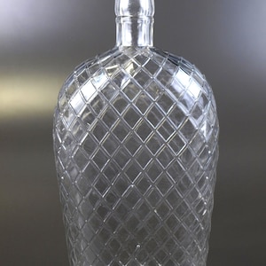Könnte beinhalten: Transparente Glasflasche mit Rautenmuster. Die Flasche hat einen abgerundeten Körper und einen zylindrischen Hals. Das Glas ist transparent und lässt Licht durch. Die Flasche ist leer und steht vor einem grauen Hintergrund.
