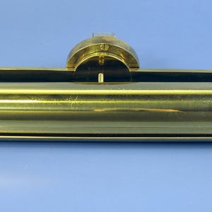 Könnte beinhalten: Ein messingfarbenes, rechteckiges Objekt mit abgerundeter Oberseite und zylindrischem Körper. Das Objekt hat eine Reihe kleiner Löcher und scheint ein Vintage- oder antikes Objekt zu sein. Der Hintergrund ist einfarbig blau.