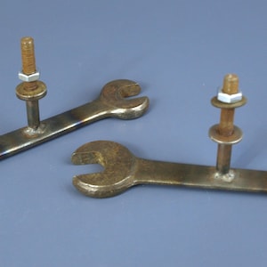 Peut inclure: Deux clés en métal, chacune dotée d'une tête ouverte aux deux extrémités, avec un boulon fileté vertical soudé au centre. Chaque boulon comprend une rondelle plate et un écrou hexagonal, présentant des signes d'oxydation et une finition sombre.