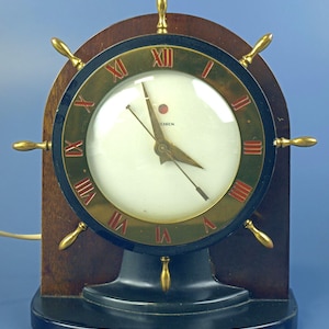 以下が含まれることがあります： 船の舵の形をしたヴィンテージ時計。時計は、濃い木製のベースと、赤いローマ数字が付いた黒と金の文字盤を備えています。時計の針は金色で、文字盤には「CHRON」と書かれています。