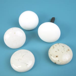 May include: Five white ceramic cabinet knobs on a blue background. The knobs vary in shape and texture, with some being smooth and round, while others have a speckled or marbled appearance. One knob has a metal screw attached.