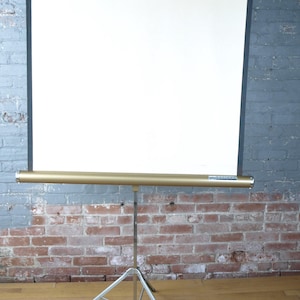 May include: A portable projection screen featuring a large, rectangular white surface with black borders. The screen is mounted on a gold-toned cylindrical housing and supported by a silver-coloured metal tripod stand with three legs.