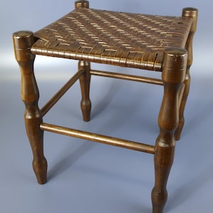 May include: A wooden stool with a woven seat. The stool has four turned legs and a simple rectangular frame.