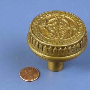 Può includere: Una maniglia per porta dorata con un designo decorativo che presenta una bussola e altri simboli. La maniglia è circa delle dimensioni di una moneta da un centesimo di euro.