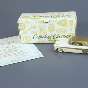 May include: A white and gold die-cast model car with a matching box and certificate. The car has a gold roof and trim, with white body panels. The box is labeled "Collector's Classics". The certificate is in English.