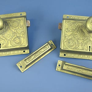 Peut inclure: Deux anciens loquets de porte en laiton avec des boutons assortis et deux plaques rectangulaires. Les loquets et les plaques présentent un motif floral orné. La quincaillerie est posée sur un fond bleu.