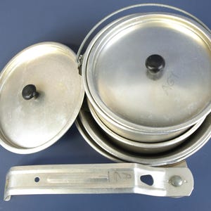 May include: A vintage aluminum camping mess kit with a stack of nesting pots, lids, and a handle. The lids have black knobs. The kit includes a metal handle and is set against a blue background.