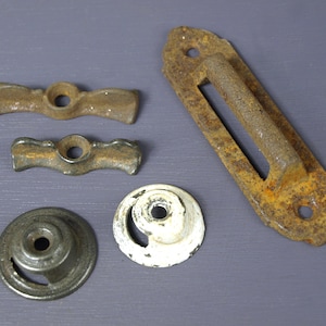 May include: Five antique metal door hardware pieces. Two are rusty, brown, and black, with a wavy shape. One is a rusty, brown, and black rectangular piece with a handle. Two are round, one is white with a black center, and the other is black with a silver center.