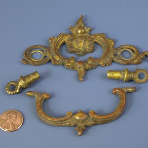 以下が含まれることがあります： 真鍮製のアンティーク家具用金具セット。装飾的なバックプレート、ハンドル、2つの小さなノブが含まれています。これらのパーツは、温かみのある金色の色合いで、装飾的で古びたデザインです。青い背景に配置されています。サイズの目安としてペニーが含まれています。