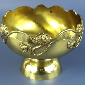 Puede incluir: Un cuenco de latón pulido con un borde festoneado y una base de pedestal. El exterior presenta motivos florales en relieve que envuelven los lados, creando un patrón decorativo continuo. La superficie metálica tiene un acabado amarillo dorado reflectante.