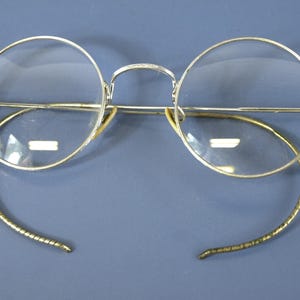 Può includere: Un paio di occhiali in stile vintage con lenti rotonde e trasparenti incorniciate in un sottile metallo color argento lucido. Gli occhiali presentano un ponte alto e aste flessibili a cavo avvolto che si curvano verso il basso.