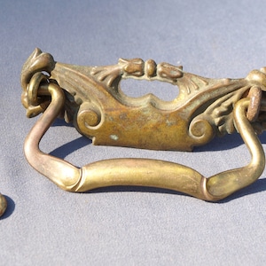 May include: Brass door pull with ornate details and a curved handle. The pull is attached to a screw and a ring.