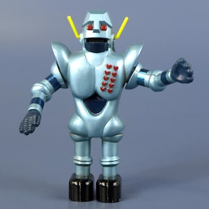 Puede incluir: Un juguete de robot azul metálico con ojos rojos, antenas amarillas y una serie de acentos rojos en forma de corazón en el pecho. El robot tiene brazos y piernas articulados, con pies negros y detalles a rayas azules y plateadas.