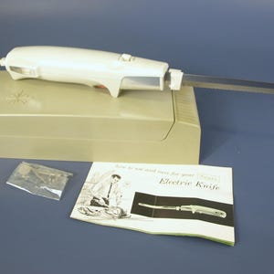 May include: An off-white electric knife with a silver blade rests on a beige base. A user manual titled "Electric Knife" and a small plastic bag are also present. The knife has a cord.