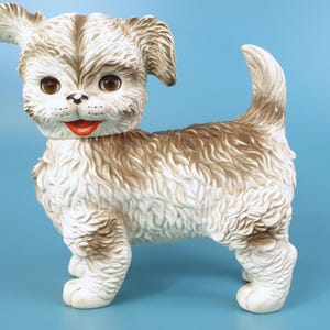Peut inclure: Figurine de chiot vintage en céramique, crème et marron. Le chiot a de grands yeux bruns, une bouche rouge et un pelage touffu. La figurine est debout sur ses quatre pattes, la queue levée.