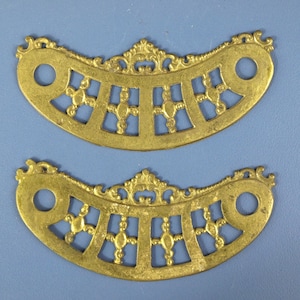 以下が含まれることがあります： 真鍮色のアンティーク装飾金属部品2点。各部品は、対称的なパターンと円形の穴を備えた、装飾的で透かし彫りのデザインの湾曲した形状をしています。部品は青色の背景にあります。