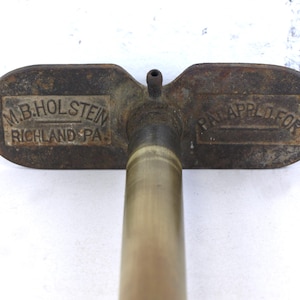May include: A vintage metal tool with a long cylindrical shaft and a wide, flat handle. The handle features embossed text reading M.B. HOLSTEIN RICHLAND PA. on the left side and PAT. APPLD. FOR on the right side, with a small nozzle at the center.