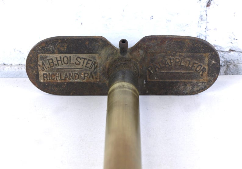 May include: A vintage metal tool with a long cylindrical shaft and a wide, flat handle. The handle features embossed text reading M.B. HOLSTEIN RICHLAND PA. on the left side and PAT. APPLD. FOR on the right side, with a small nozzle at the center.