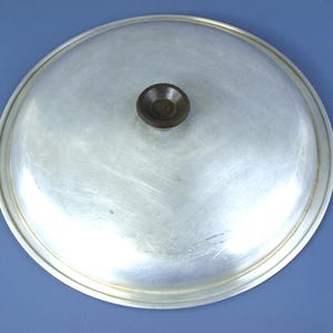 May include: A round, silver-colored metal pot lid with a smooth, slightly domed surface and a small, dark brown circular knob centered at the top. The lid features a simple, clean design with a flat rim edge, showing a metallic finish.