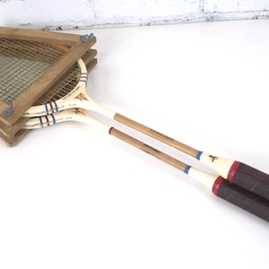 May include: Two vintage wooden tennis rackets secured within a wooden press frame. The rackets feature white frames with red and blue striped accents, natural wood shafts, and dark brown grips. The press is held together by four metal wing nuts.
