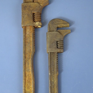 May include: Two heavy-duty metal pipe wrenches with a weathered, rusted brown finish. Each tool features an adjustable jaw mechanism with a threaded nut and a serrated handle. The smaller wrench displays embossed text along the handle shaft.