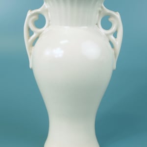 Puede incluir: Un jarrón de cerámica de color blanco roto brillante con un borde acampanado y acanalado, y dos asas simétricas caladas en forma de voluta a los lados. El cuerpo del jarrón tiene una silueta lisa y redondeada que se estrecha hacia una base.