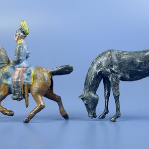 Op de afbeelding: Twee antieke speelgoedpaarden, een met een ruiter in blauw uniform en gele hoed, de andere een donkergrijs paard met zijn hoofd naar beneden. Het speelgoed heeft een versleten, verouderd uiterlijk.