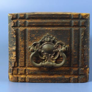 May include: A rectangular wooden drawer front with a dark brown finish and visible signs of wear along the edges. The drawer features a grid-like carved pattern and a central ornate brass drop handle with intricate floral and scroll detailing.