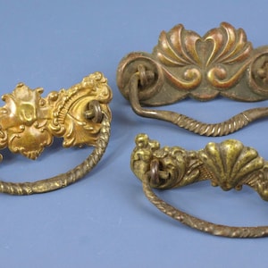 Può includere: Tre maniglie antiche per cassetti in ottone con disegni ornati. Le maniglie presentano intricati dettagli floreali e a volute, con manici curvi. L'ottone ha una calda patina invecchiata, che suggerisce l'artigianato vintage. Lo sfondo è blu.