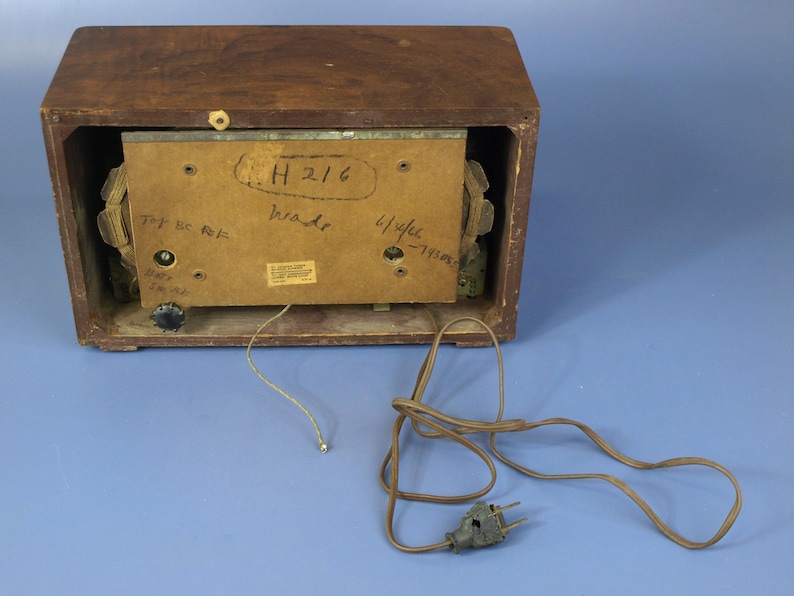 Puede incluir: La parte posterior de una radio vintage de madera con un cable marr&oacute;n y un enchufe de dos clavijas. La radio tiene una etiqueta que dice "H 216" y "114 kc - 1150 kc".