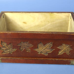 May include: A rectangular wooden box with a dark reddish-brown exterior. The box features four embossed leaf designs with a gold finish. The corners are reinforced with metal brackets. The interior is a light yellow colour.