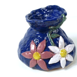 以下が含まれることがあります： テクスチャ加工された表面と3Dの花が付いた青いセラミックの花瓶。花はピンク、白、黄色です。