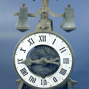 May include: An antique clock with a white face and black Roman numerals. The clock has a gear mechanism visible in the center and two bell-shaped weights on either side. The base has the Roman numeral MCDXL. The clock is made of a weathered gray metal.