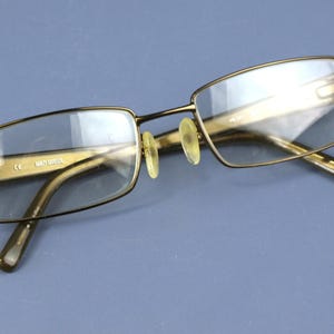 May include: Gold-framed rectangular eyeglasses with clear lenses. The glasses feature a double bridge and adjustable nose pads. The arms are a darker gold colour. The text "Gaultier" is visible on the frame.