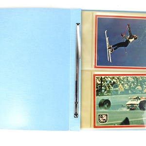 May include: A light blue binder with a silver metal spine, open to display two vintage sports trading cards. One card features a skier in mid-air, the other a race car on a track.