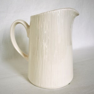 Puede incluir: Una jarra de cerámica blanca con una superficie texturizada y un asa. La jarra está diseñada con líneas verticales.