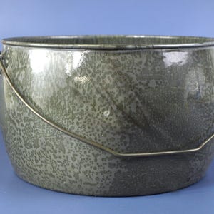 May include: A round, gray-green ceramic pot with a speckled pattern and a metal handle. The pot has a wide opening and a curved metal handle for carrying. The exterior has a glossy finish.