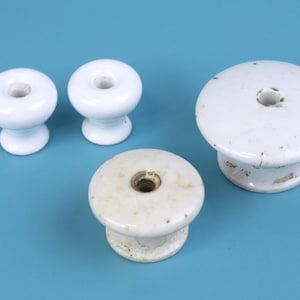 以下が含まれることがあります： 光沢のある白いセラミックキャビネットノブ4個。2つのノブは小さくて丸く、他の2つは平らな上面を持つ大きめのノブです。ノブは無地の青い背景に配置されています。