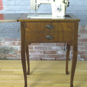 May include: A vintage sewing machine with a cream-colored body and silver accents sits atop a wooden cabinet. The cabinet has two drawers with brass-colored pulls and slender, curved legs. The machine is ready for sewing.
