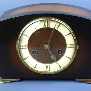 May include: A vintage wooden mantel clock with a curved top and a round clock face. The clock face has Roman numerals and gold-colored hands. The clock is brown with a lighter wood grain on top.
