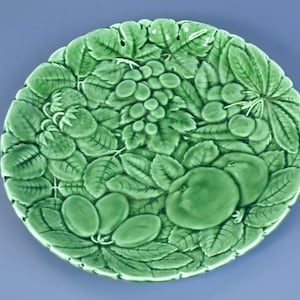 以下が含まれることがあります： さまざまな果物と葉のレリーフデザインが施された緑色のセラミックプレート。 プレートには、ブドウ、イチゴ、サクランボ、プラム、リンゴが描かれています。 プレートは丸く、詳細で質感のある表面を持っています。