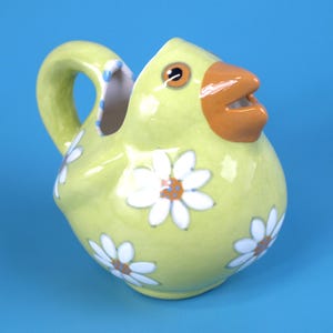 Puede incluir: Jarra de cer&aacute;mica caprichosa con forma de pollito amarillo con pico naranja y ojos marrones. Dise&ntilde;os de flores de margarita blancas est&aacute;n dispersos por el cuerpo. El asa es curva y el interior es blanco con lunares azules.