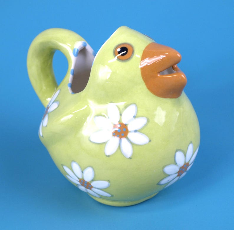 Puede incluir: Jarra de cer&aacute;mica caprichosa con forma de pollito amarillo con pico naranja y ojos marrones. Dise&ntilde;os de flores de margarita blancas est&aacute;n dispersos por el cuerpo. El asa es curva y el interior es blanco con lunares azules.