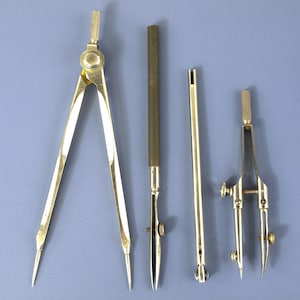 May include: A set of four gold-toned metal drafting tools including a large compass, a ruling pen with a dark handle, a straight metal rod, and a smaller precision compass with adjustable screws and two pointed tips for technical drawing.