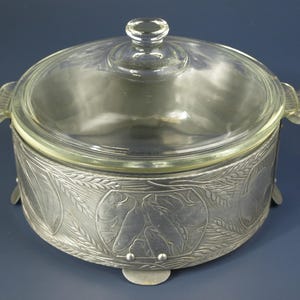May include: Silver-toned metal serving dish with a glass lid. The dish has a decorative design of wheat and fish. The dish has four feet.